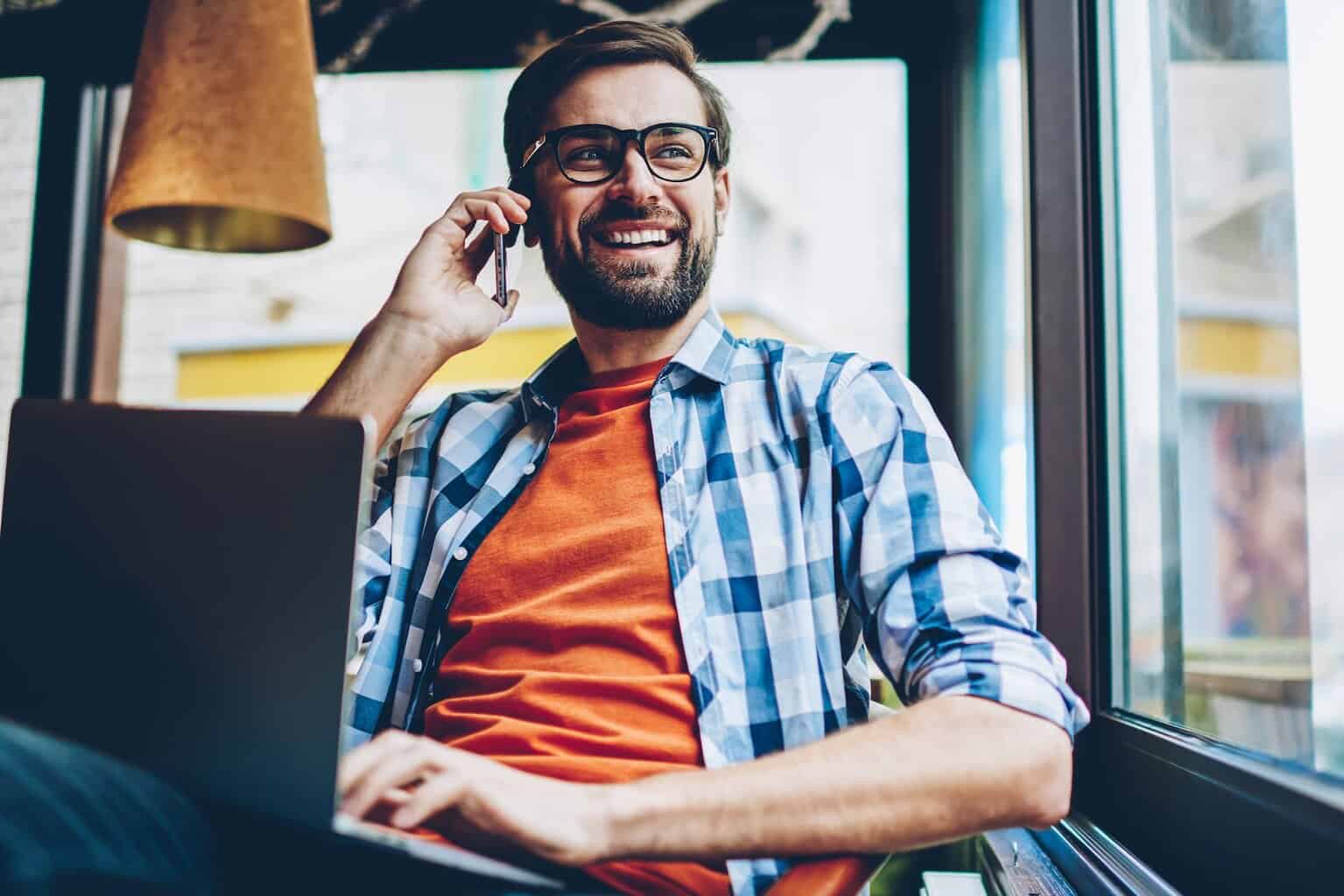 A man with glasses and a beard sitting at a table with a laptop and talking on the phone